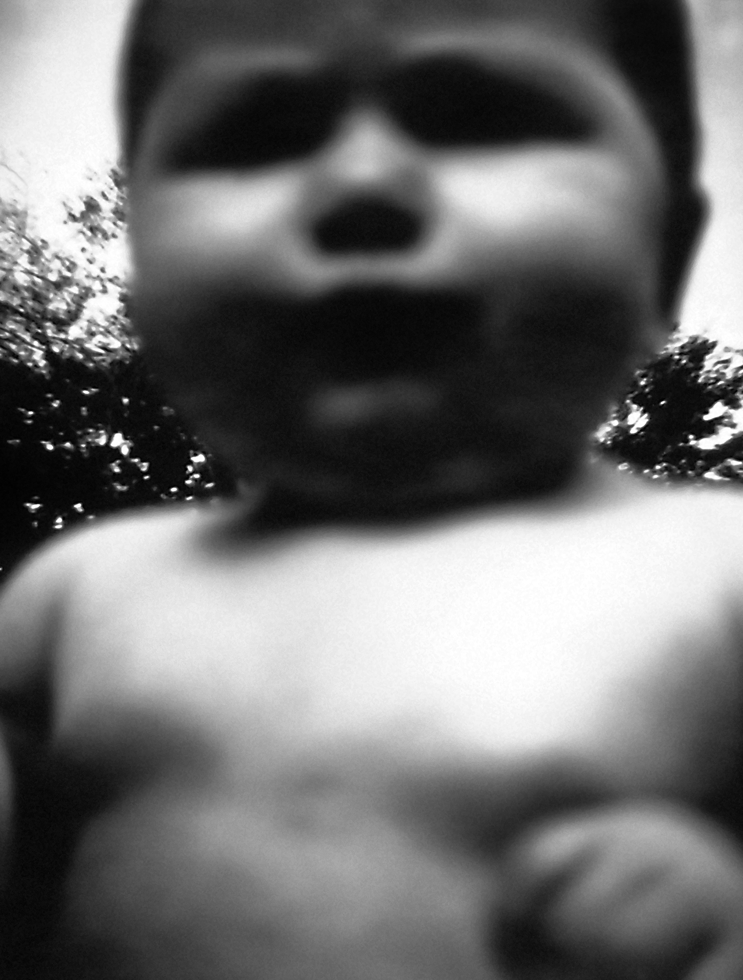 Close-up of the nude upper half of a toddler standing in front of bushes. The toddler’s image is blurry and the toddler appears inordinately large.