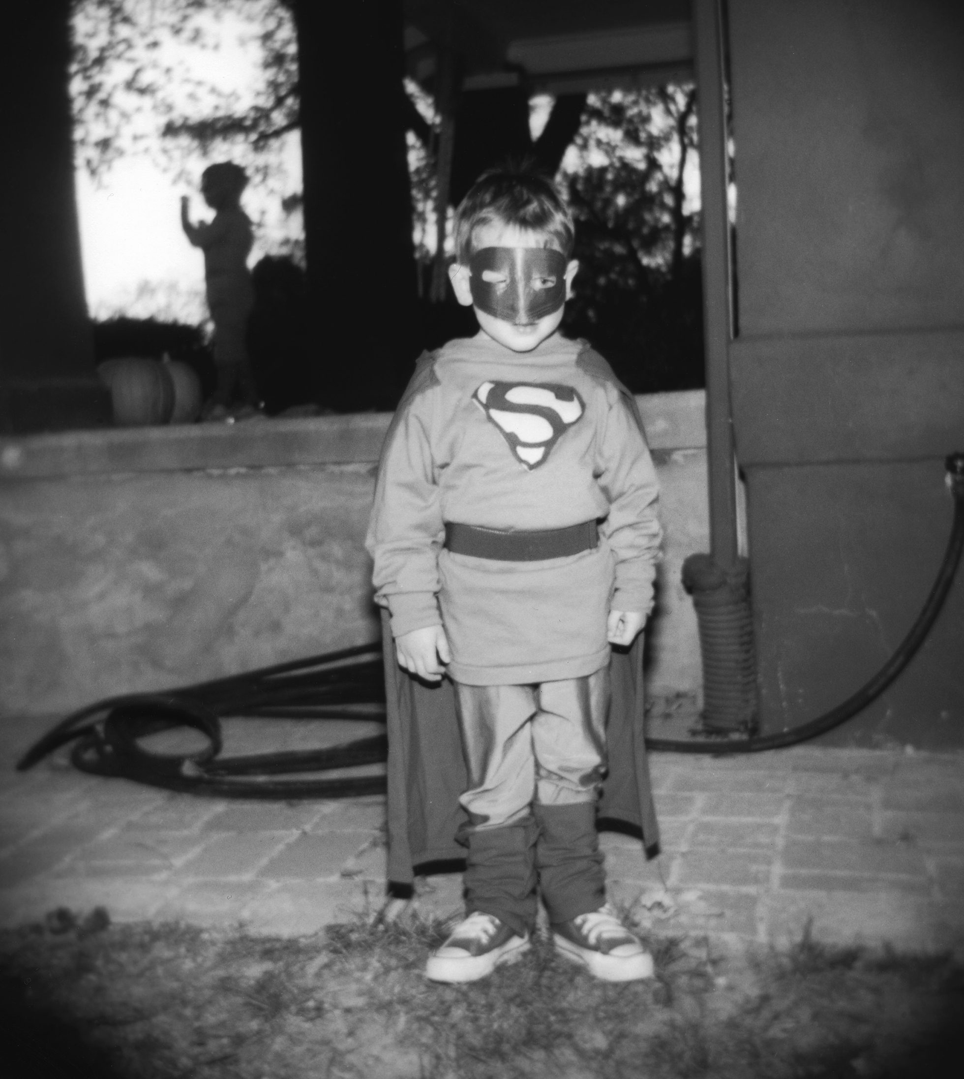 A young boy in a Superman costume with cape and mask stands facing the camera near the patio of a house.