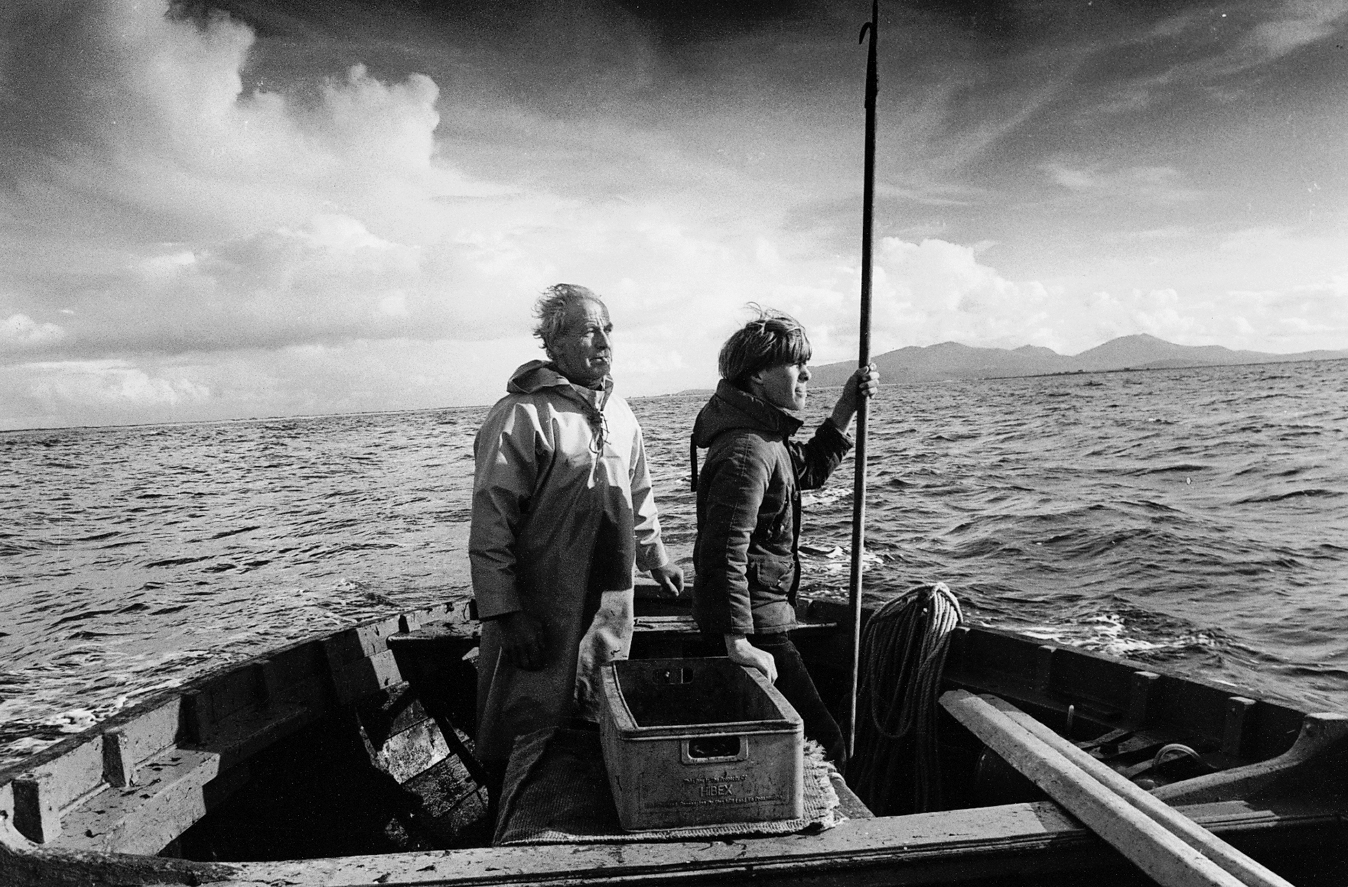 An old man and a young man both in profile looking off to the right in a nondescript boat off the Outer Hebrides, Scotland, on a choppy sea on a cold, cloudy day. The young man is holding a long-handled harpoon pointed up in his left hand.