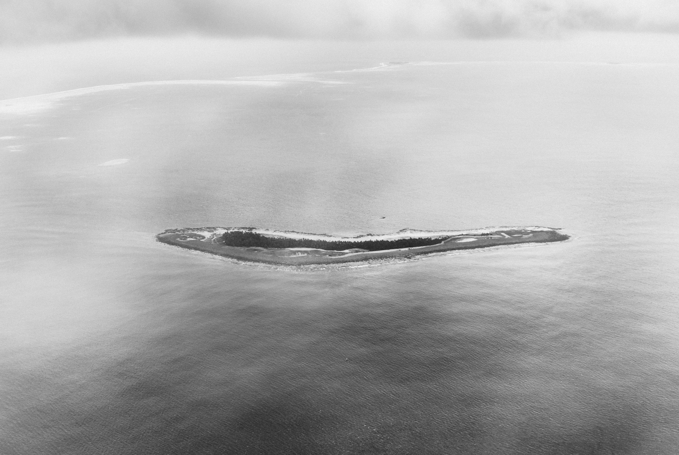 A long, narrow island surrounded by water and sky that appear white. The whiteness enveloping the strip of land evokes a haunting vulnerability.