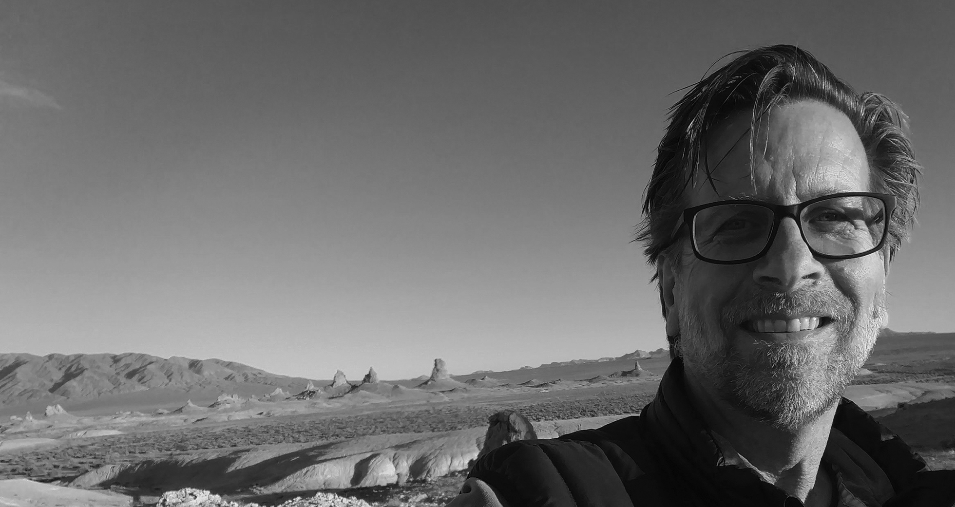 Douglas Christie has a mustache and beard and is wearing glasses as he smiles amongst the desert setting behind him.