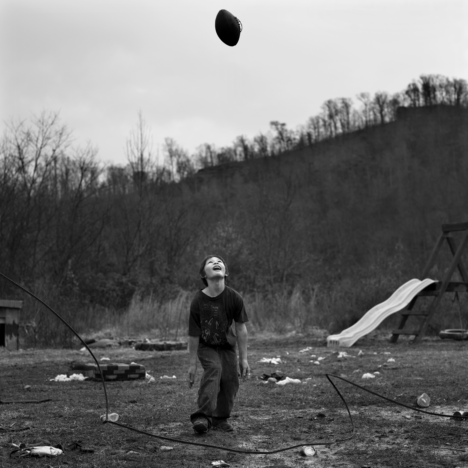 A boy looks up at a football with its laces coming apart in the air above him as he stands in a trash-filled yard with a slide and a tire swing to the right. The yard is in front of a hill with bare-branched trees.