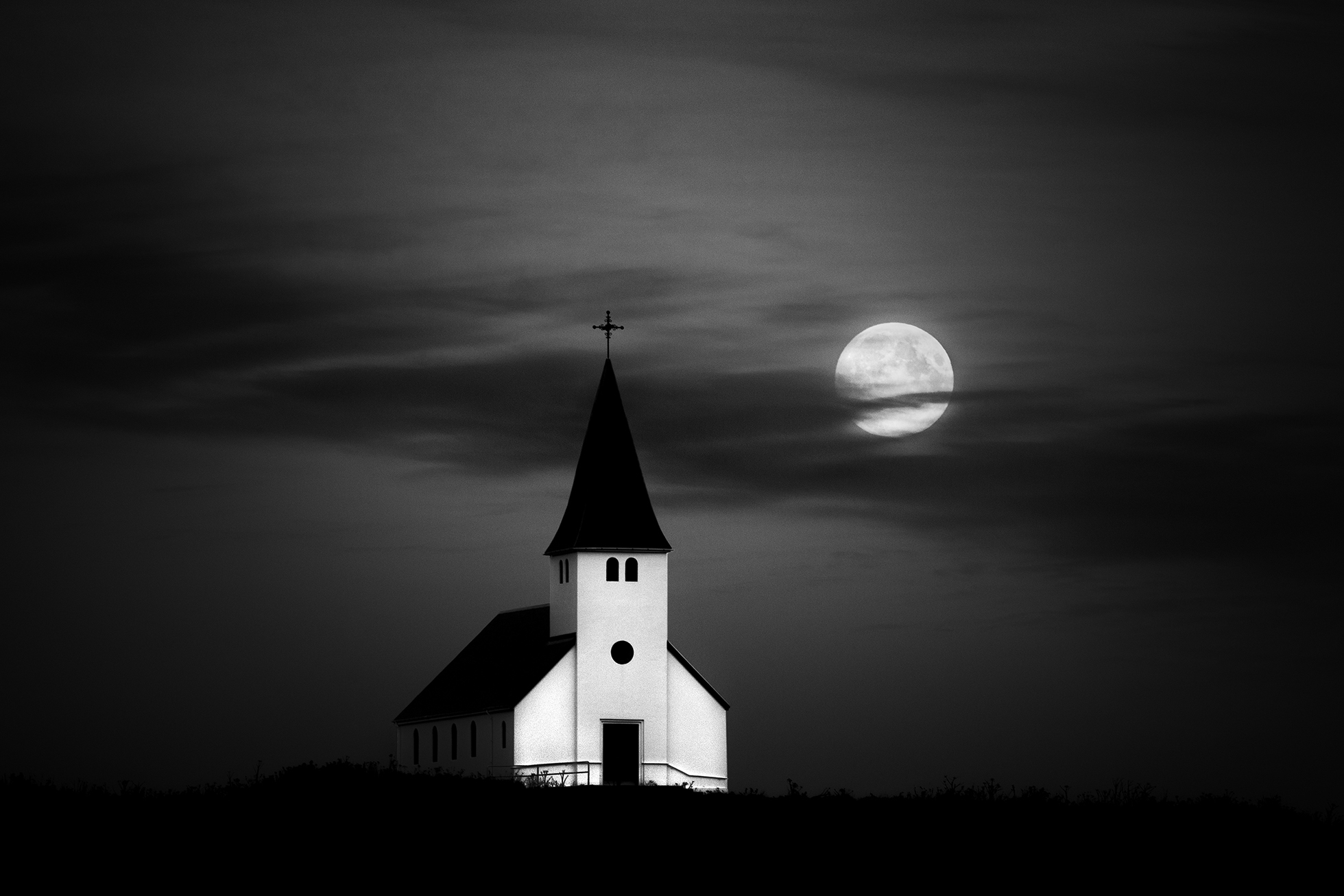 A clean-lined, white with black roof, single-spired church in a rural setting on a night with a prominent full moon.