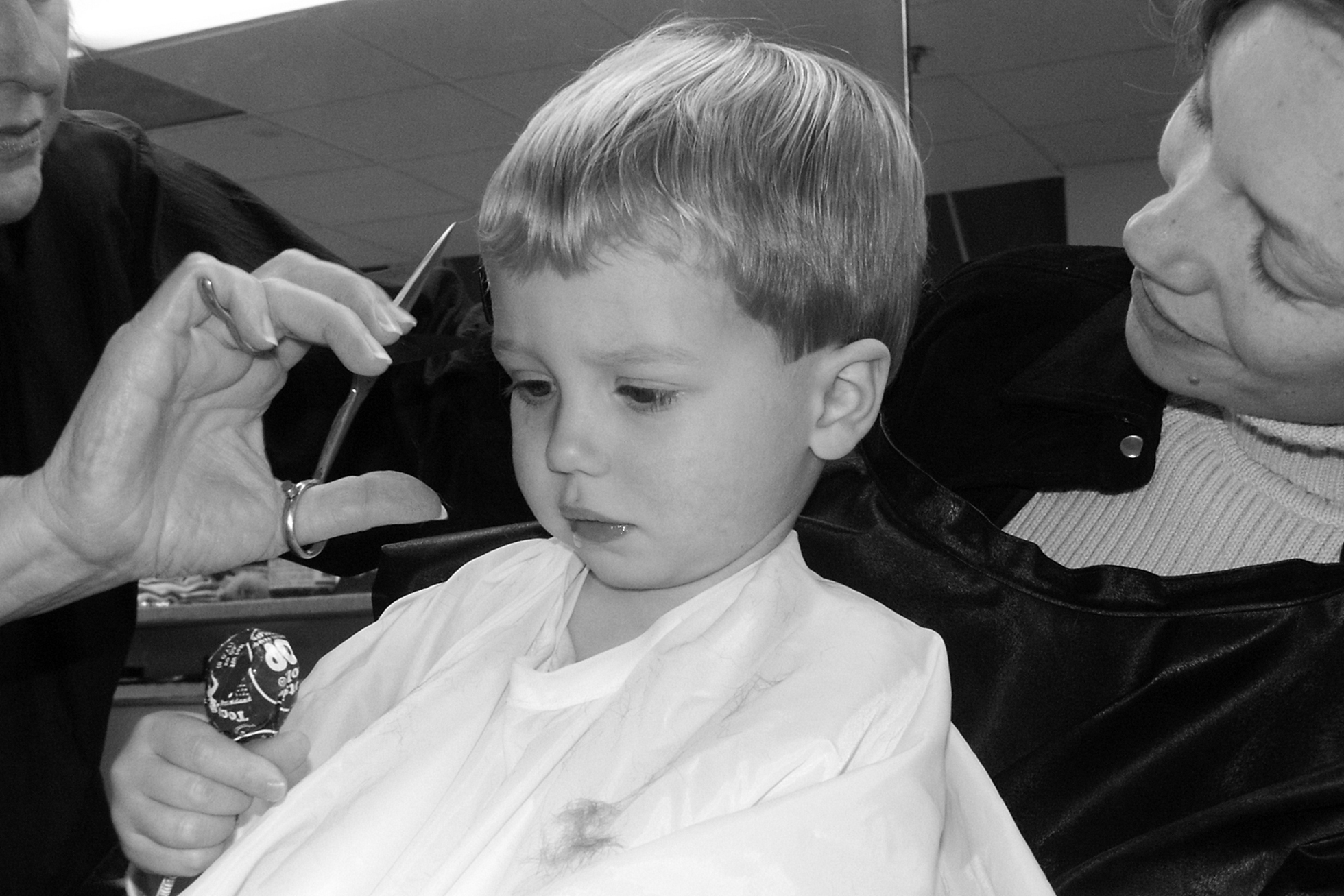 A small boy in a white hair-salon cape is sitting on a woman’s lap while getting a haircut. The boy is looking down at the Tootsie Pop in his hand.