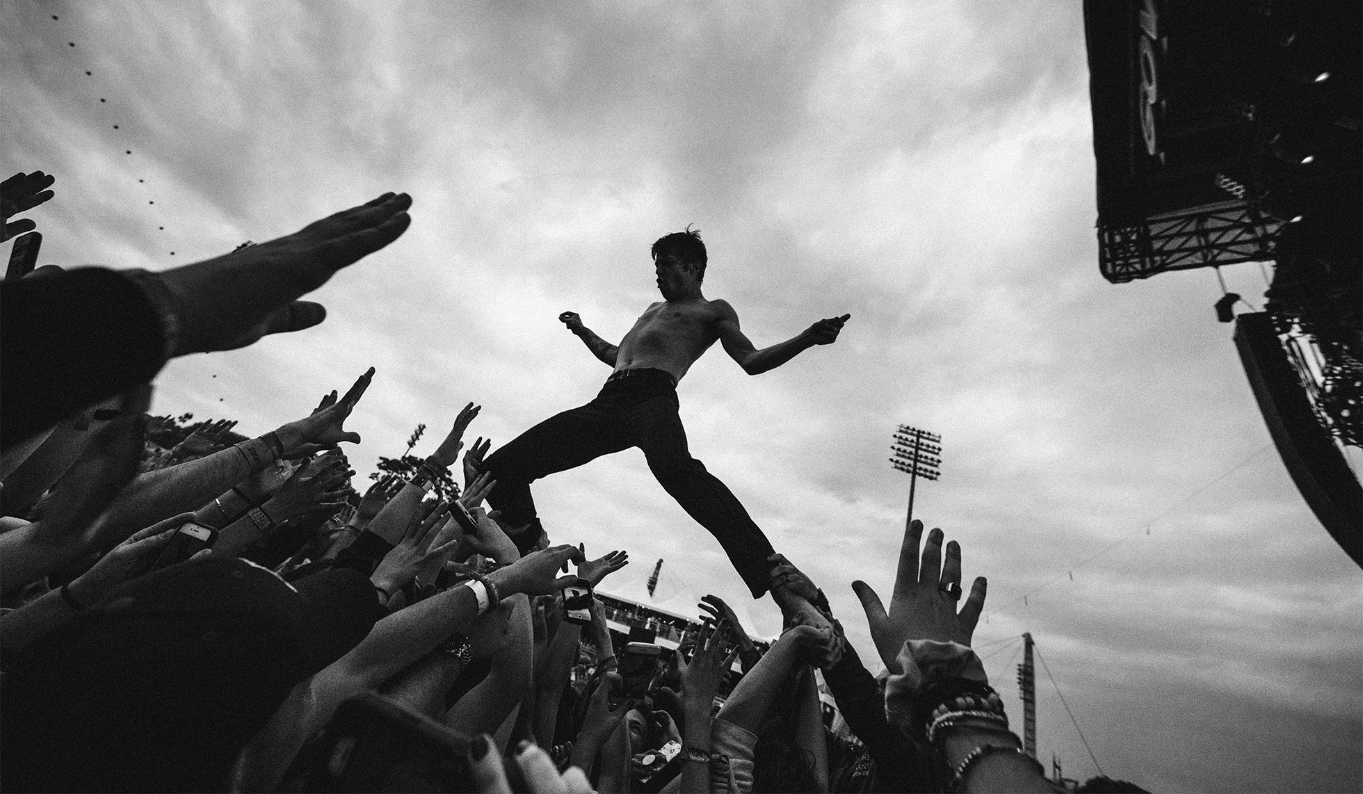 A bare-chested and barefooted young man in jeans stands on top of a crowd with his arms out to the sides at an outside event. His feet and ankles are supported by the offered hands of the crowd below.