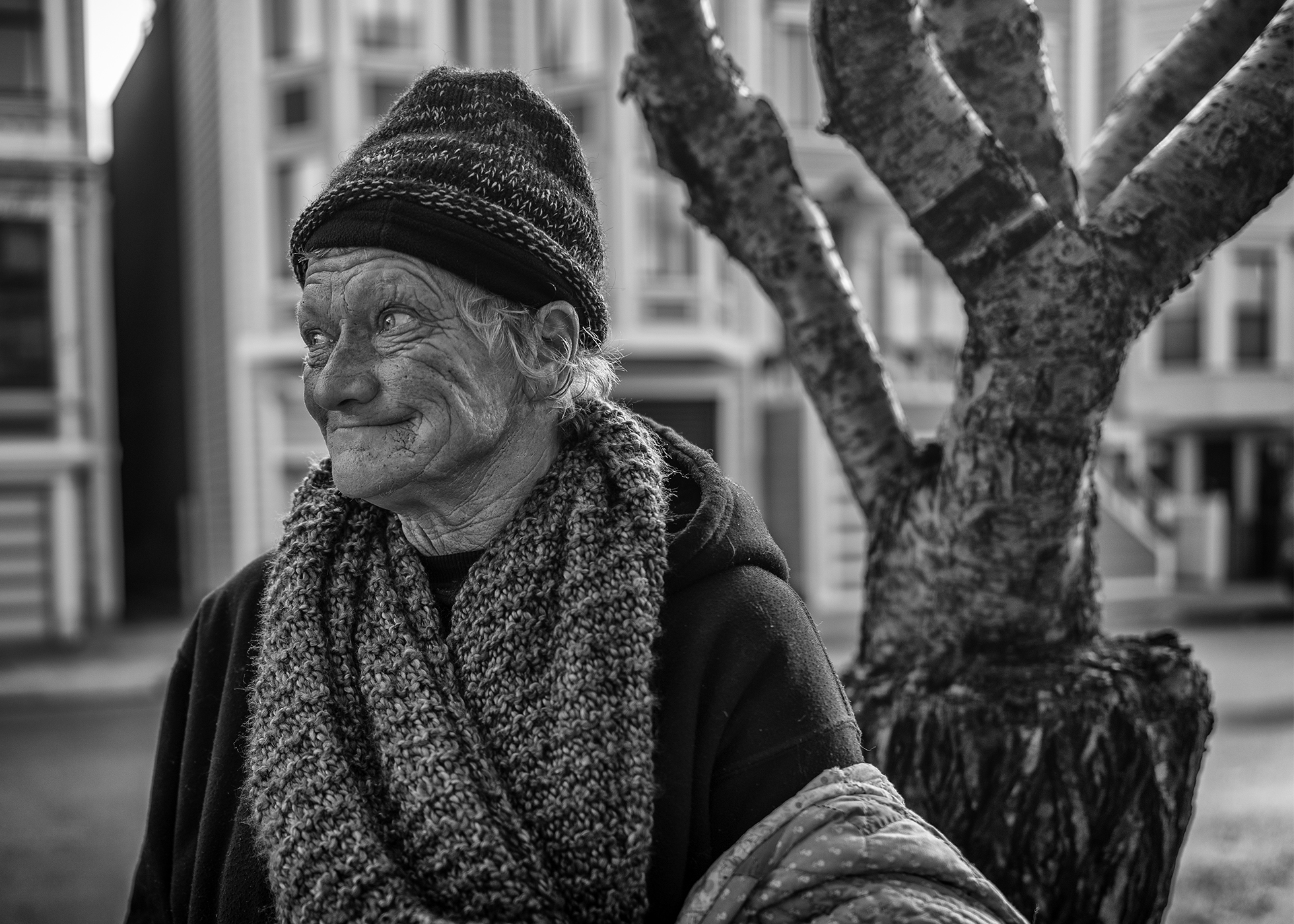 Jonalyn looks off to the side and smiles with her mouth closed. She stands in front of a tree with buildings in the background in San Francisco in December 2020. She is wearing a wool cap, winter scarf, and a hoodie.