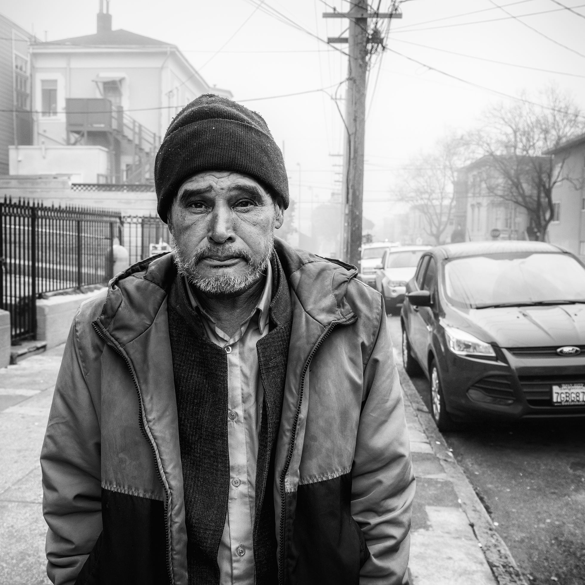 Mario looks into the camera and stands on a residential sidewalk with cars parked along the street. He is wearing a winter cap and jacket in San Francisco in January 2020.