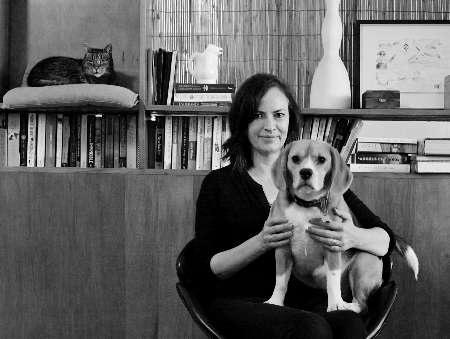 Rebecca Priestley sits in front of a bookcase with a dog on her lap, as a cat on a pillow on a shelf in the bookcase looks on.