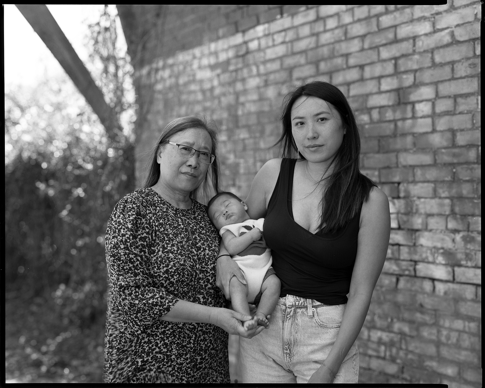 WeiWua stands next to FeiFei who holds Willow, who is asleep, against her hip with her right arm. WeiWua and FeiFei look into the camera while they stand in front of the side of a brick building in Savannah, Georgia.