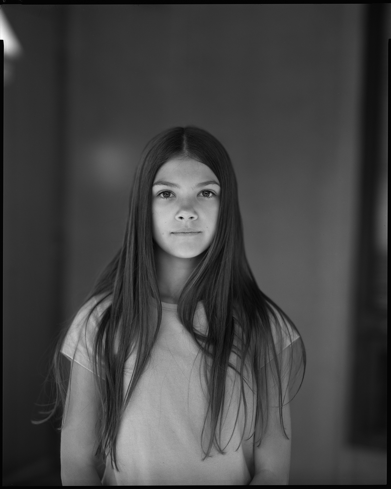 Izzy stands in front of an interior wall in Green Mountain Falls, Colorado. She has long, straight hair that flows around her shoulders and down to her waist. Izzy looks into the camera and she wears a short-sleeve shirt.