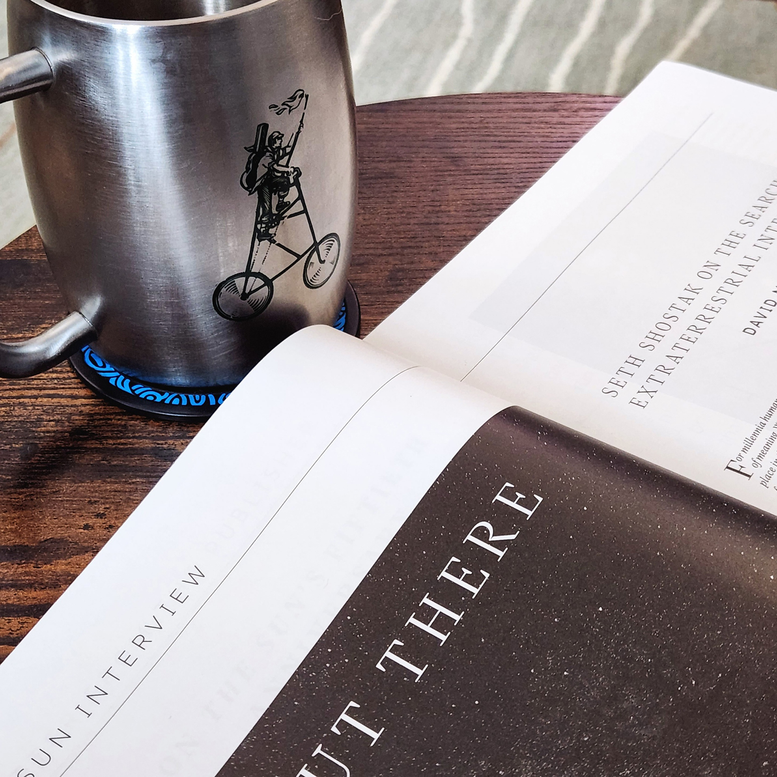 A silver mug on a table with the January 2023 issue of The Sun open to the interview Out There, Seth Shostak interviewed by David Mahaffey. On the mug is a  nineteenth-century lamplighter bearing a guitar case and a torch while riding a tall bike.