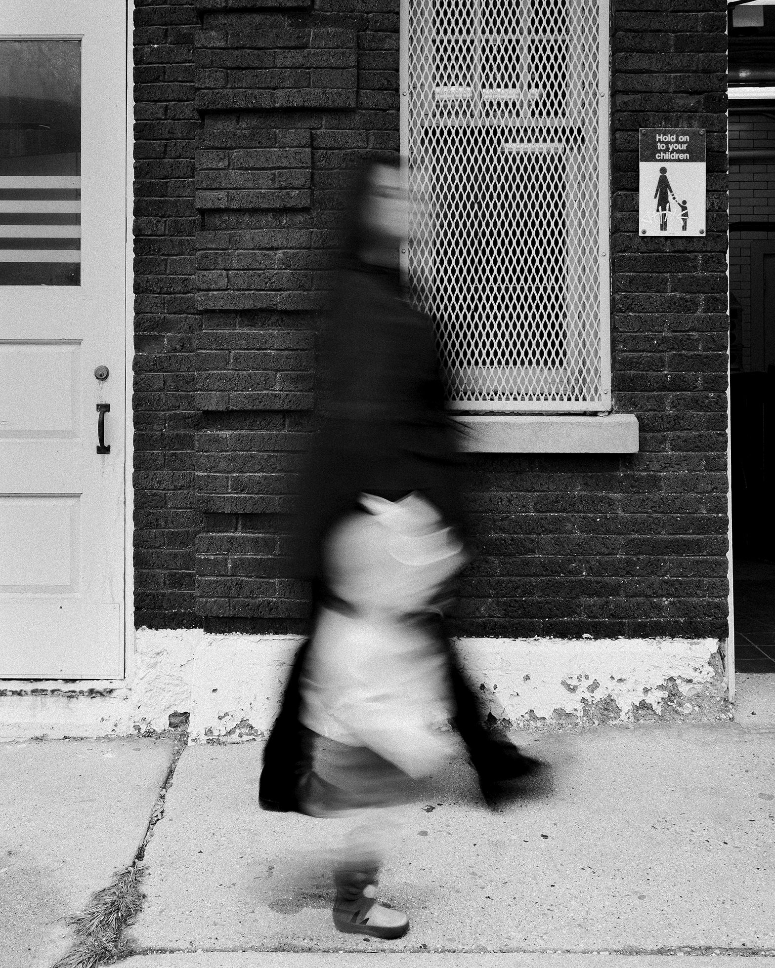 A woman and a child walk on a sidewalk past a brick building with a sign that reads “Hold on to your children.” The woman and child are motion-blurred against an intact background