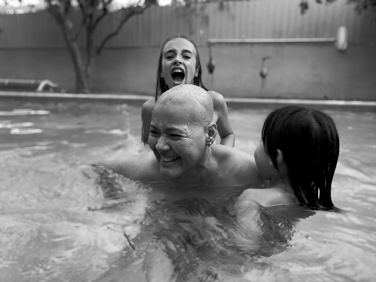A joyful, bald-headed woman smiles brightly as she plays in an above-ground pool with two young girls.