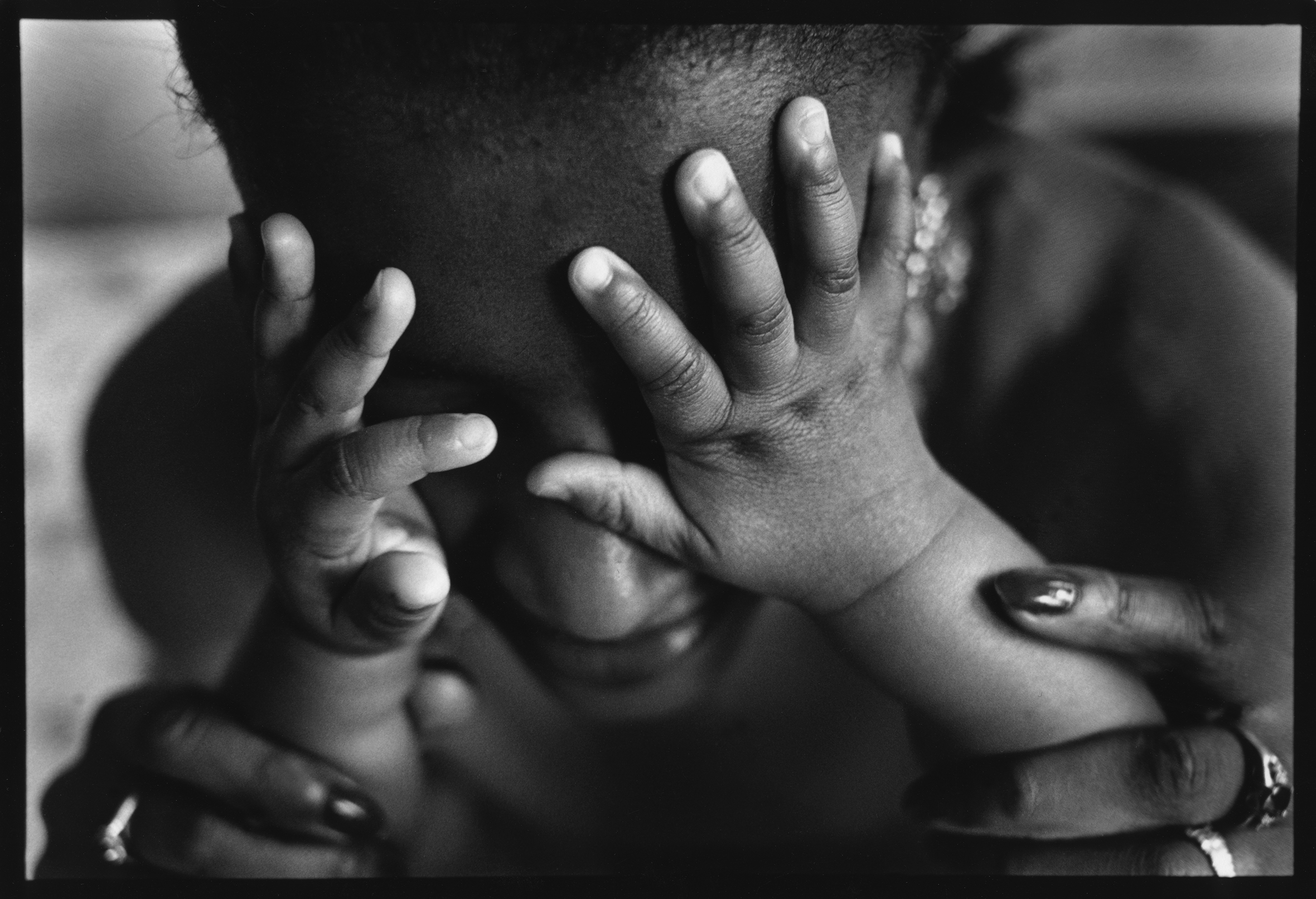 Close-up of an infant’s hands on a woman’s face obscuring her eyes. The woman is looking down on the infant and she has her hands around the infant’s forearms.