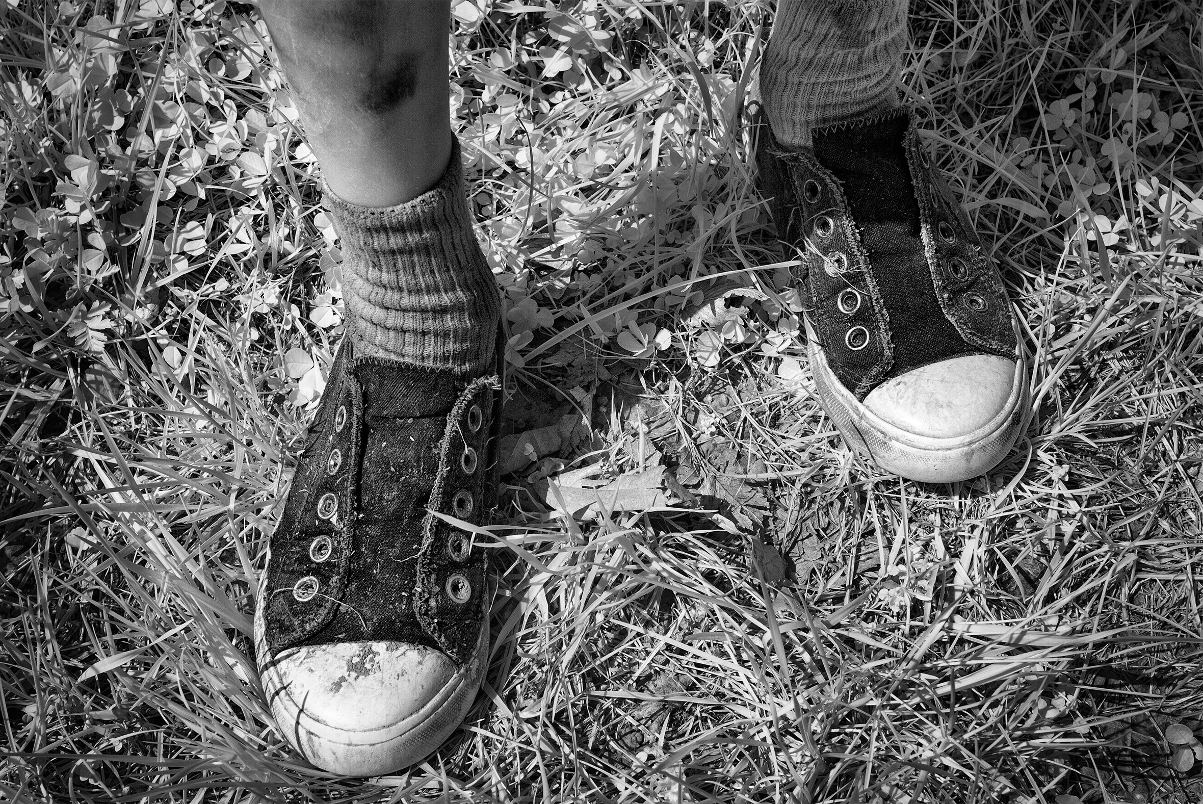 A young boy’s legs, midcalf down, smudged with dirt, with the focus on the short socks and dirty, worn, old-style tennis shoes with no laces.