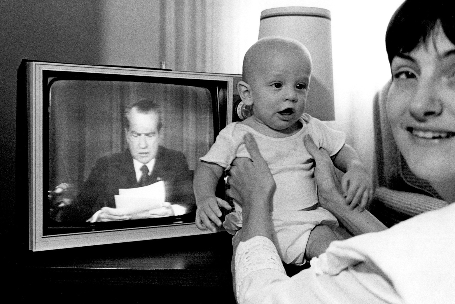 A young mother turns to look at the camera while holding her infant son up to the TV. It is the evening of August 8, 1974, and the TV in the background shows President Nixon as he addresses the nation announcing his intention to resign.