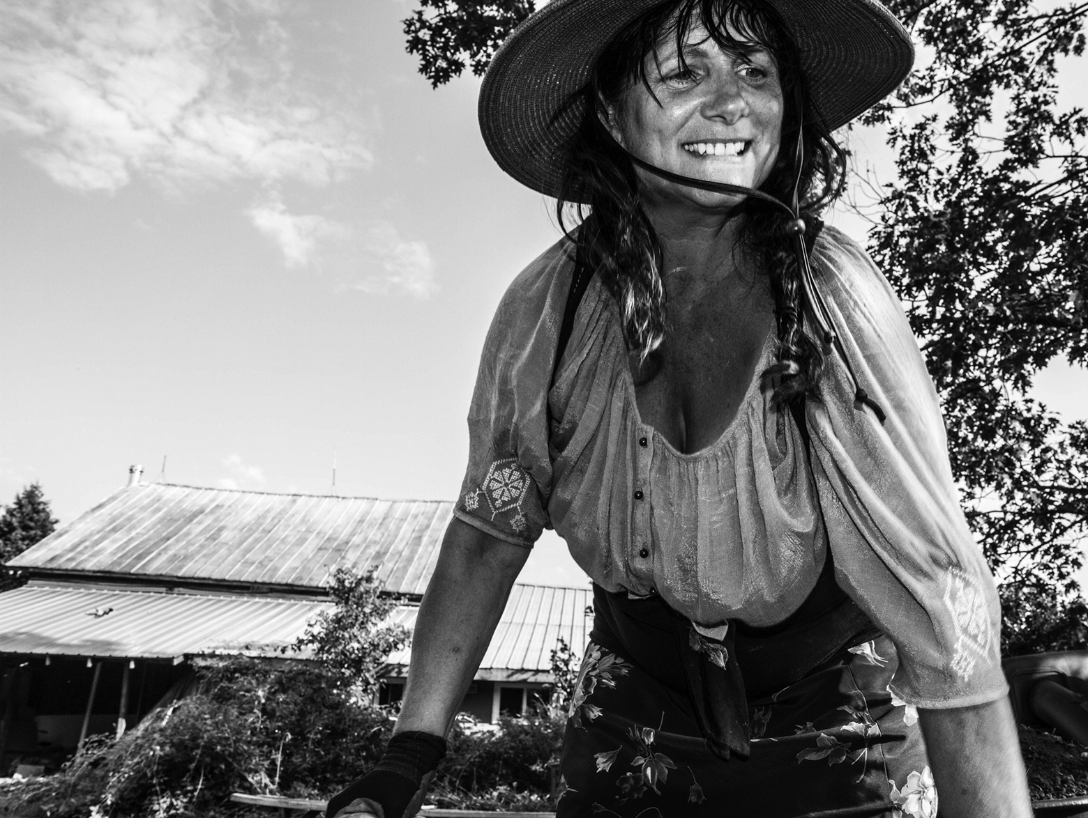 A smiling woman wearing an embroidered peasant blouse, hat, and work gloves is handling a wheelbarrow on her farmland in rural northern New York.