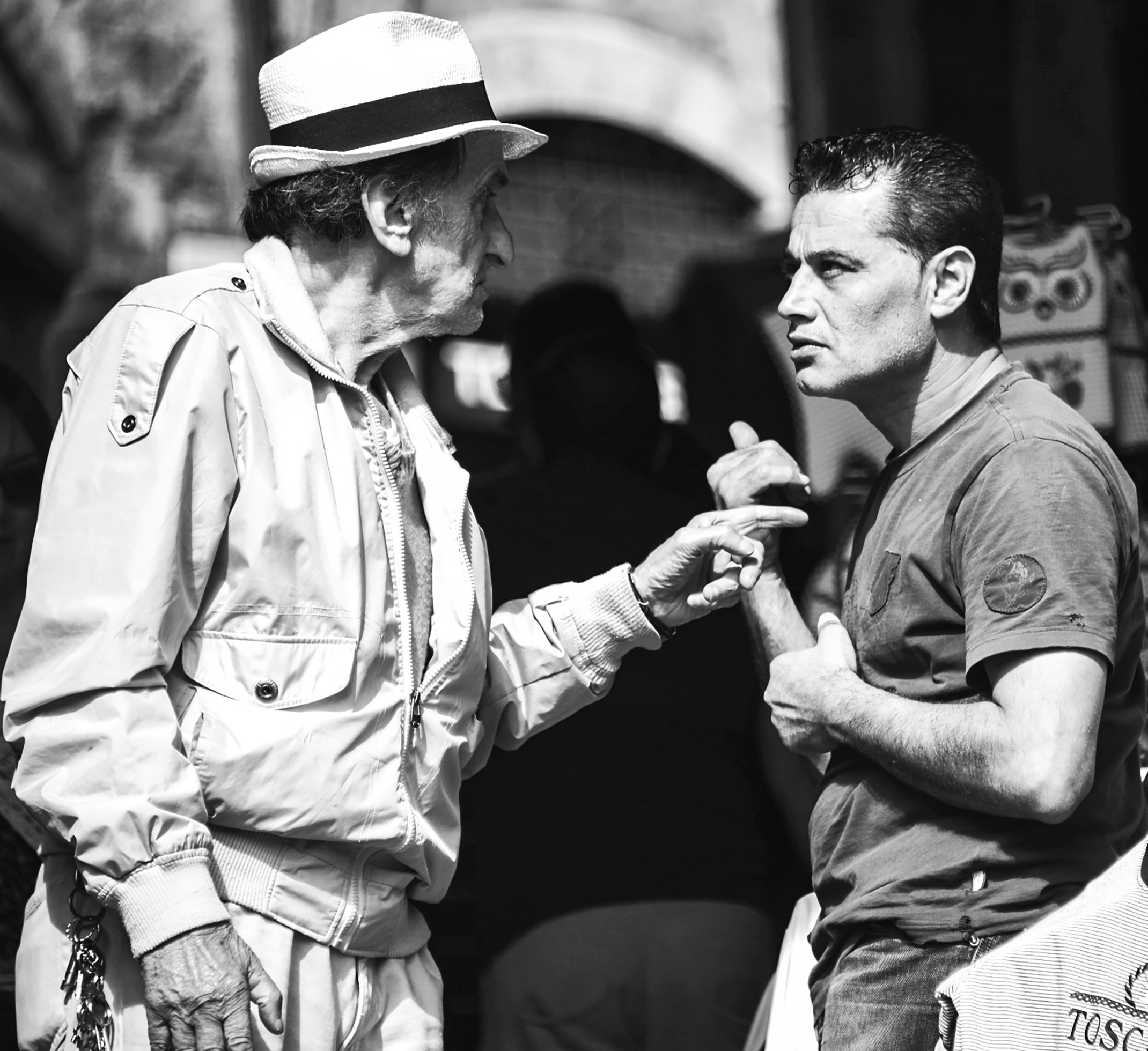 Two men stand looking intently at each other. The old man in the hat points his finger at the other man, almost touching his chest.