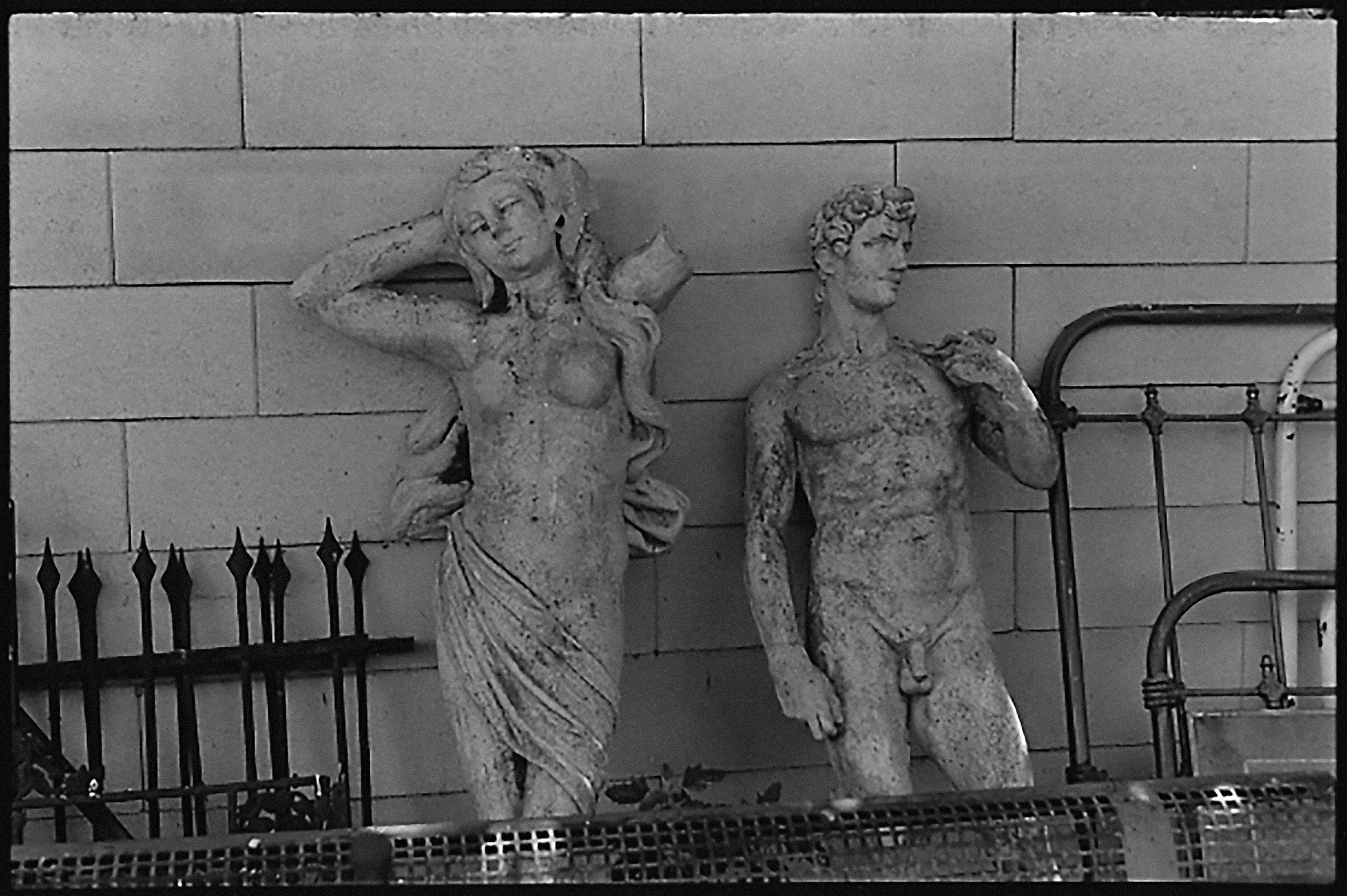 Two statues propped against a wall in a storage area amid decorative metal fencing. A statue of a mostly nude female on the left and a replica of Michelangelo’s David on the right.